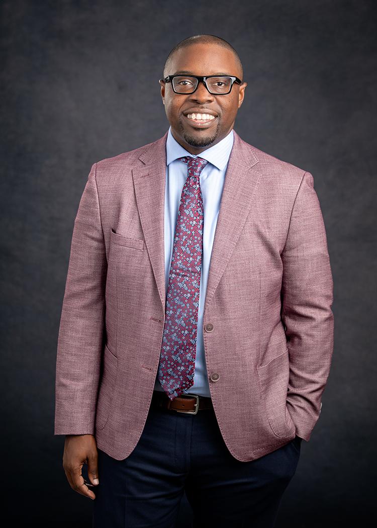 Dr. Jermaine Whirl, a smiling African American male, wearing a light red suit jacket, light blue collared shirt, and black pants with a red and blue dotted tie smiles at the camera wearing black rimmed glasses pictured against a grey background.